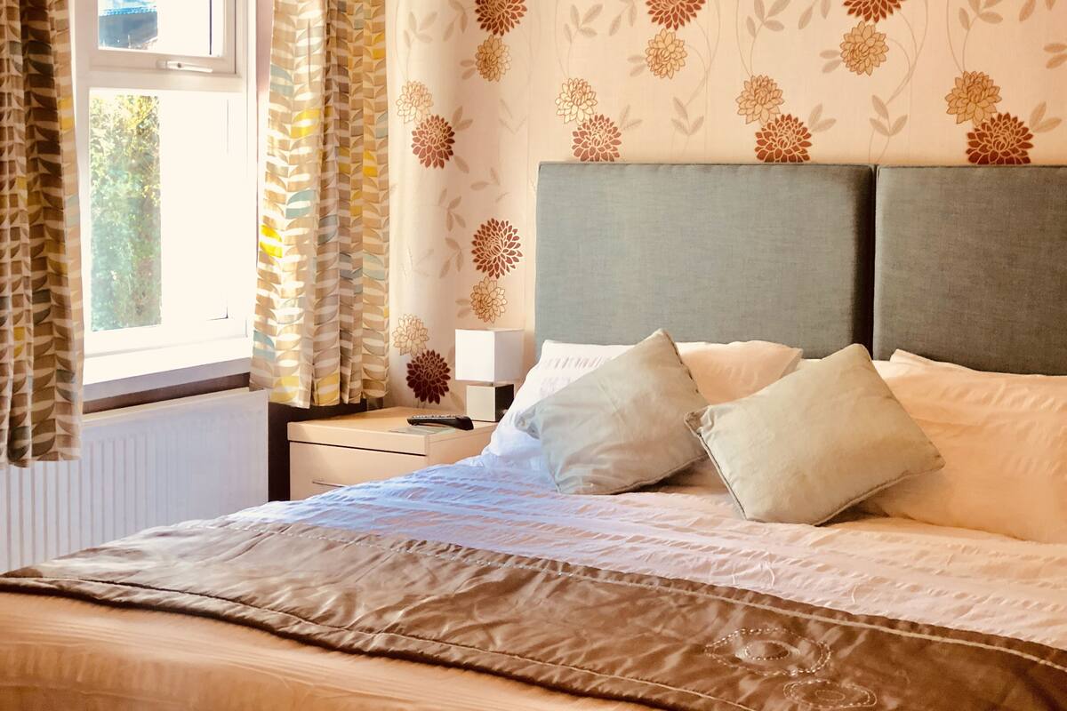 A bedroom with a bed, pillows, a nightstand, a lamp, and a window with curtains.