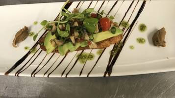 A white plate with a sandwich topped with greens, tomatoes, and a drizzle of sauce.