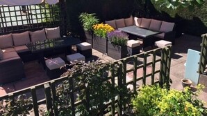 Een terras buiten met een loungeset, een tafel en stoelen, omringd door een hek en potplanten.