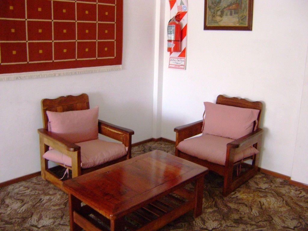 Una habitación con dos sillones de madera con cojines rosados, una mesa de centro de madera, una alfombra estampada, un tapiz blanco y rojo a rayas y un cuadro en la pared.