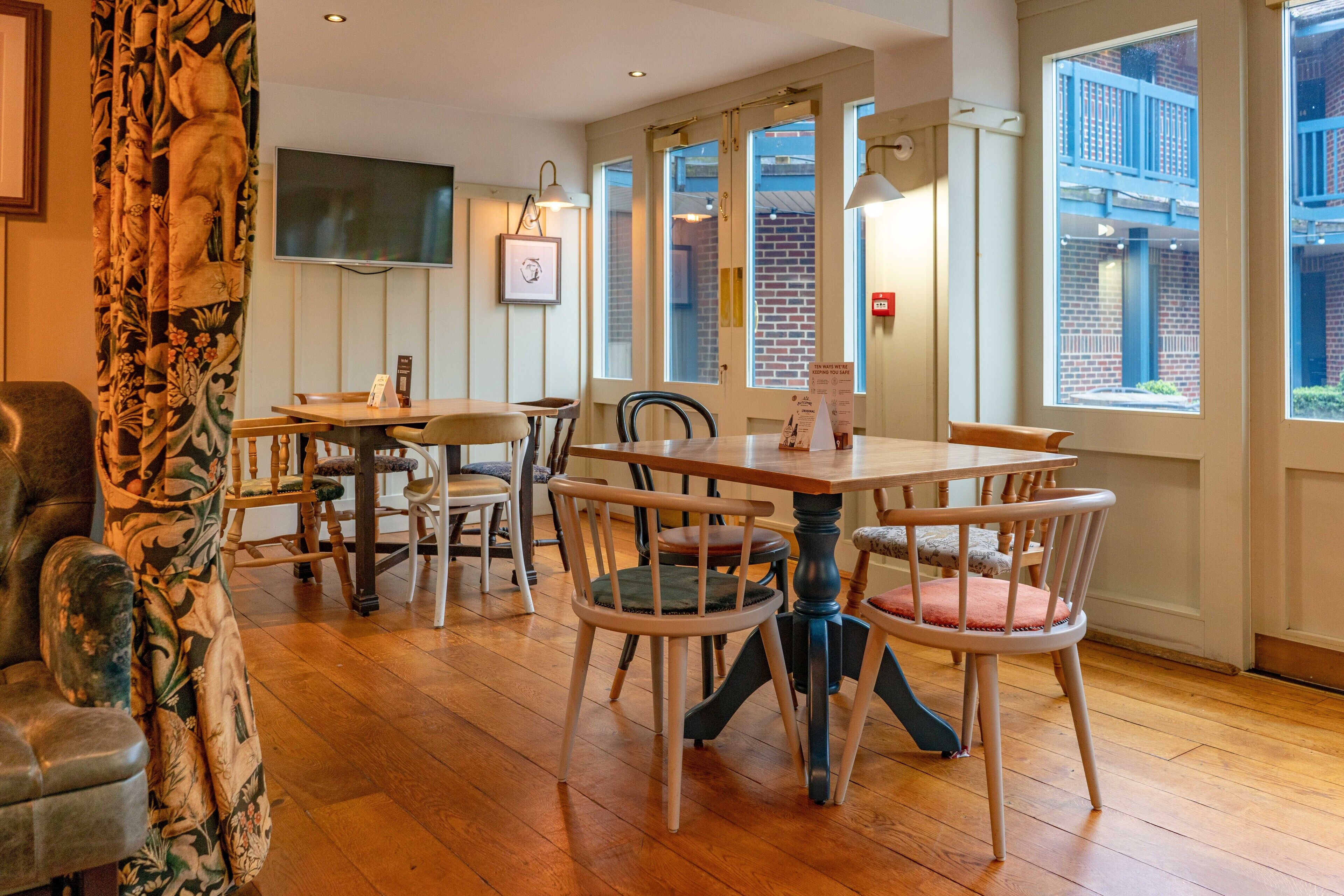 A dining area with a wooden table and chairs, a television mounted on the wall, and large windows with a view of a brick building.