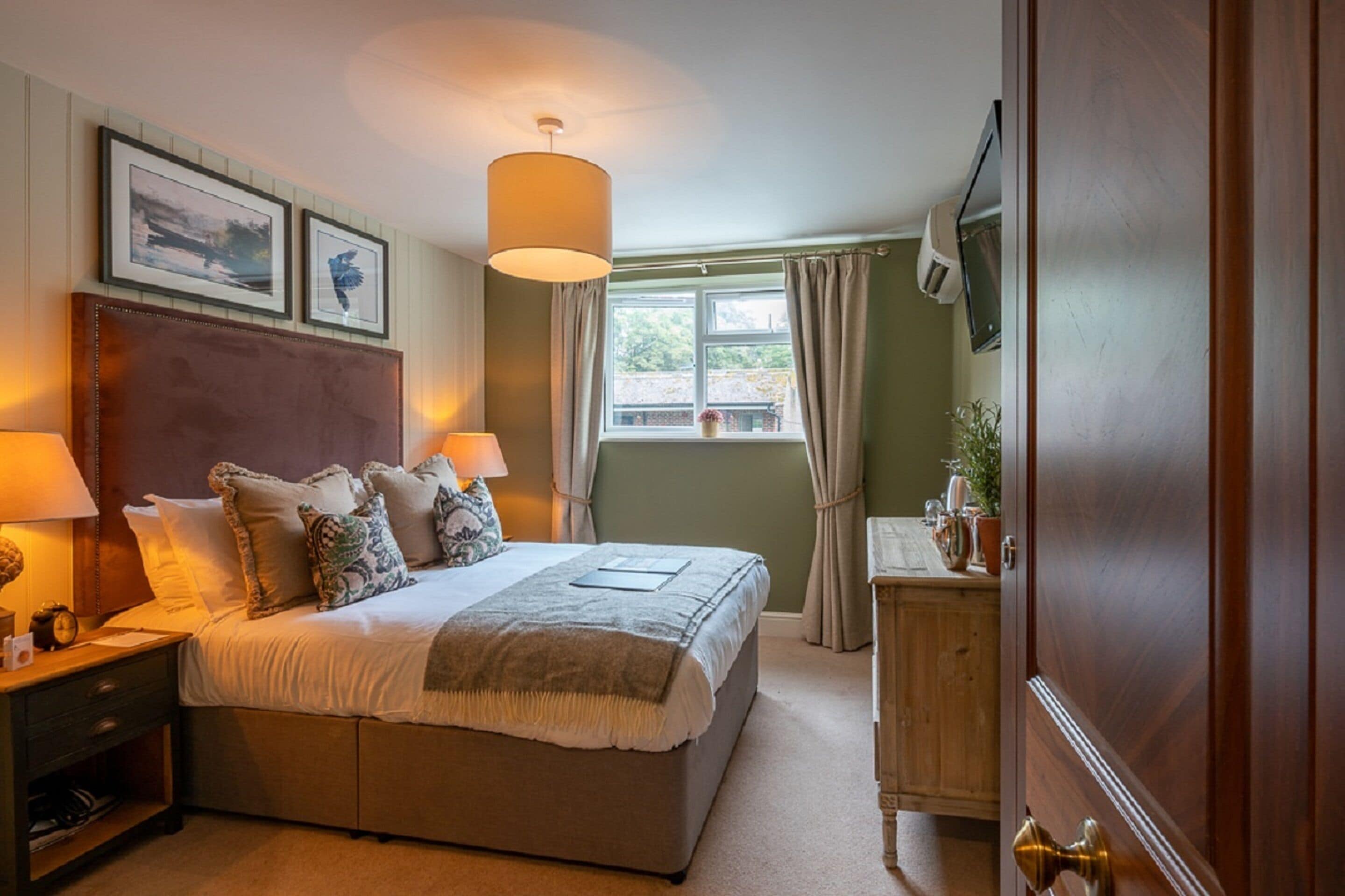 Une chambre avec un grand lit, des lampes de chevet, une table de chevet en bois, une commode en bois, une fenêtre avec des rideaux et des tableaux encadrés accrochés au mur.