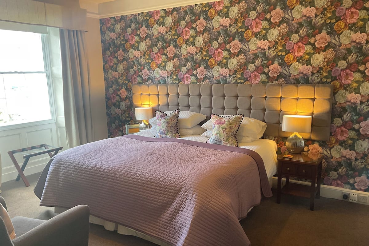 A bedroom with a floral wallpaper, a tufted headboard, two bedside lamps, and a large bed with a pink quilt.