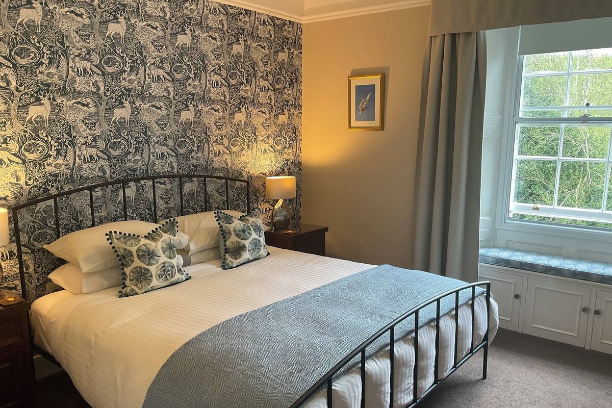 A bedroom with a patterned wallpaper, a bed with a white and blue bedspread, and a window with a view of trees.