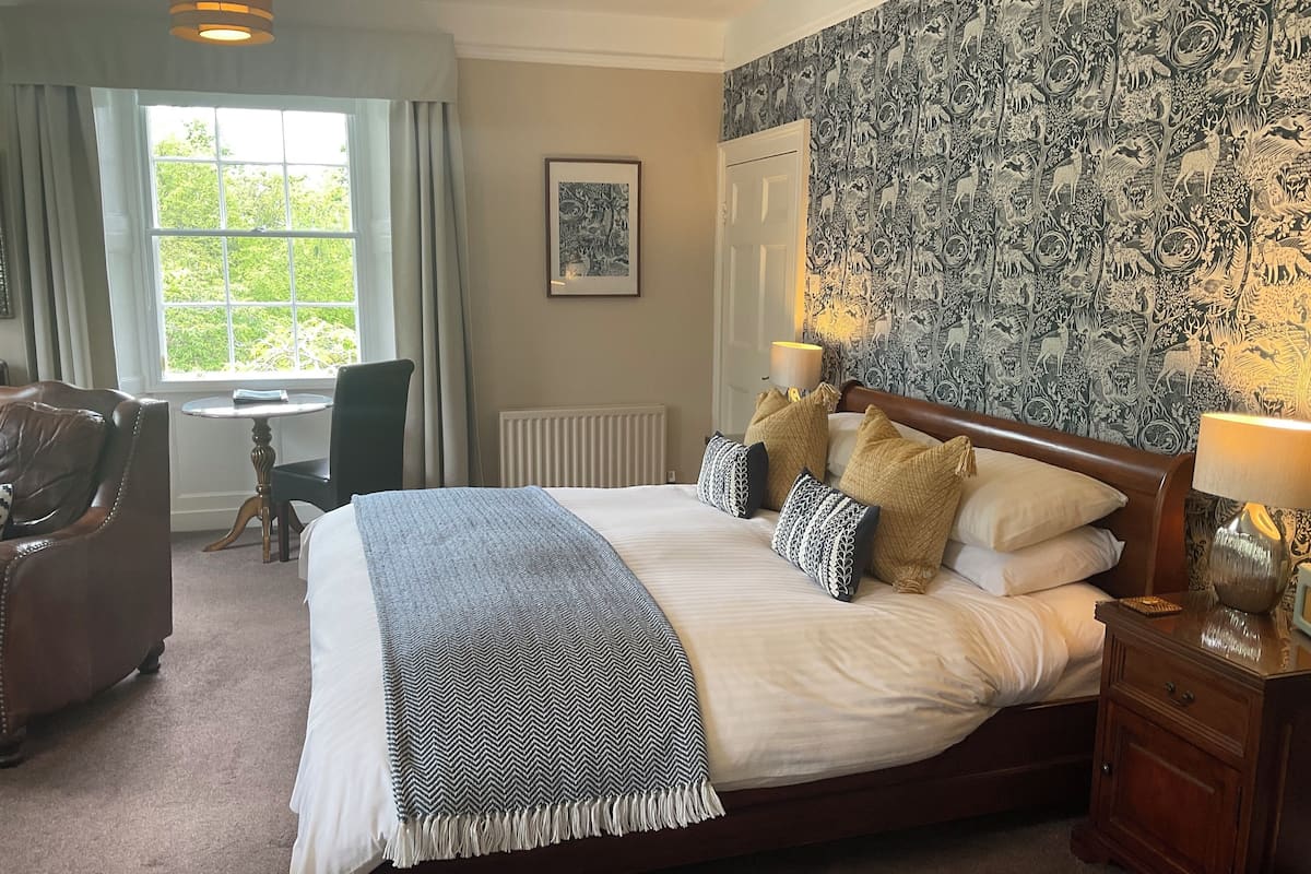 A neatly made bed with a patterned throw, a bedside lamp, and a wall with a nature-themed wallpaper.