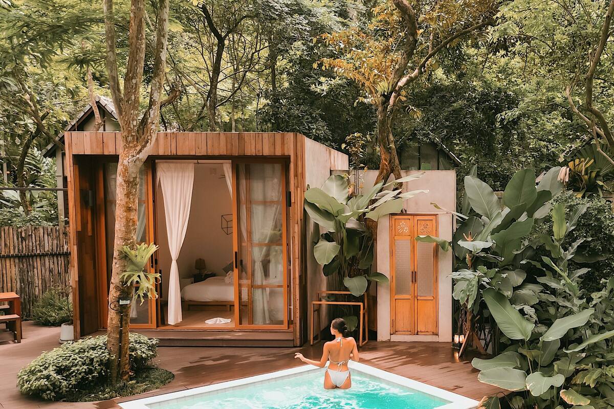 Una cabaña de madera con puerta de vidrio, una piscina pequeña y abundante vegetación alrededor.