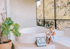 A child sitting on steps next to a bathtub, with a potted plant and a laundry basket nearby.