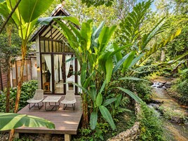 Una terraza de madera con dos sillas y una mesa, rodeada de exuberante vegetación y un pequeño arroyo.