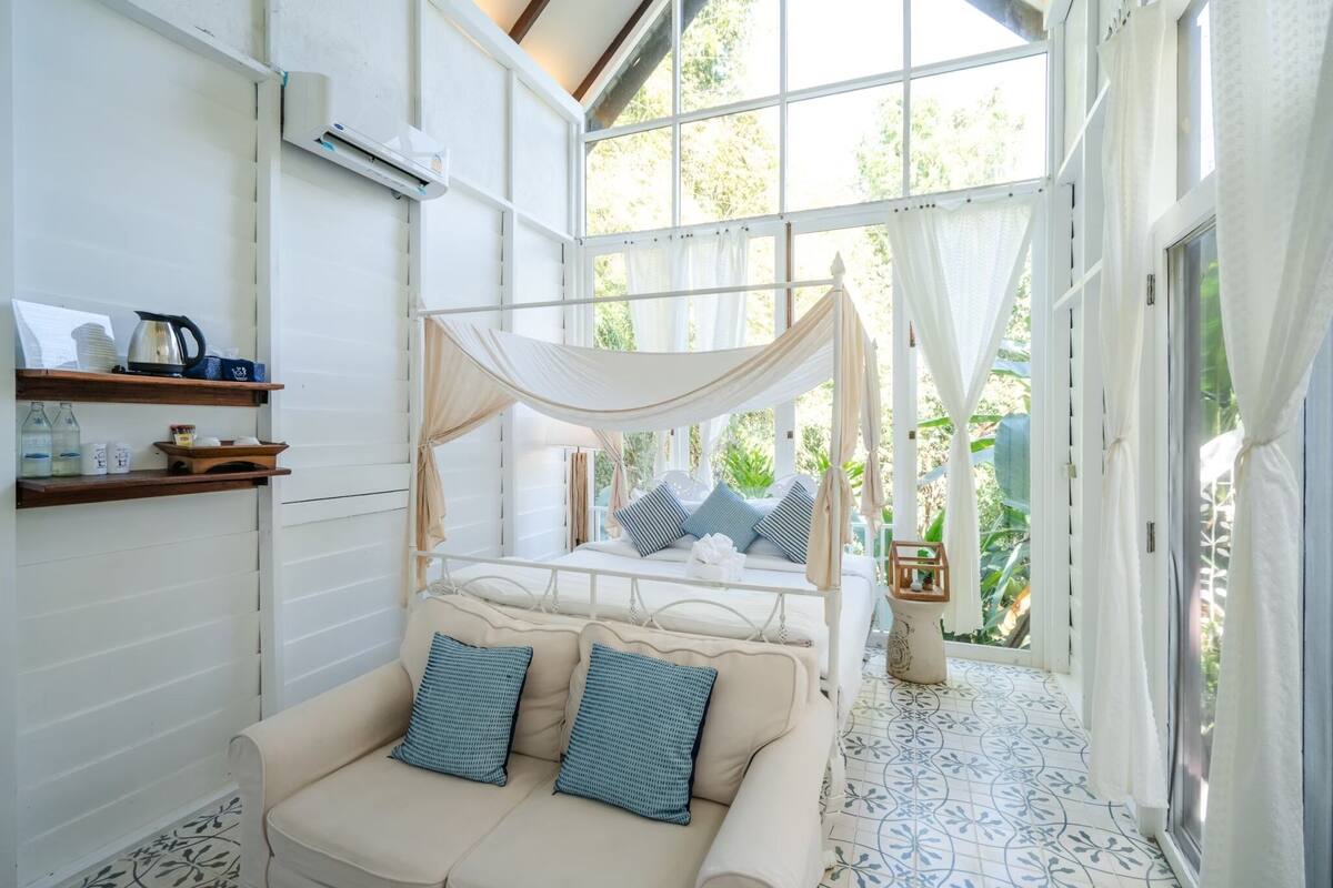 Un dormitorio con cama con dosel, un sofá con cojines, una mesita de noche y una ventana con cortinas.