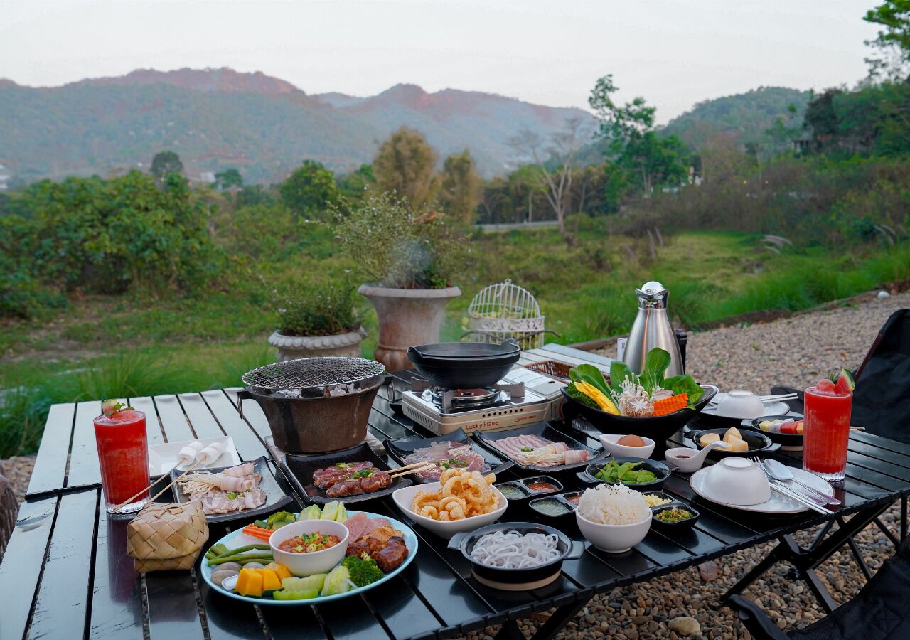 An outdoor dining table with various dishes, a grill, and a scenic mountain view in the background.