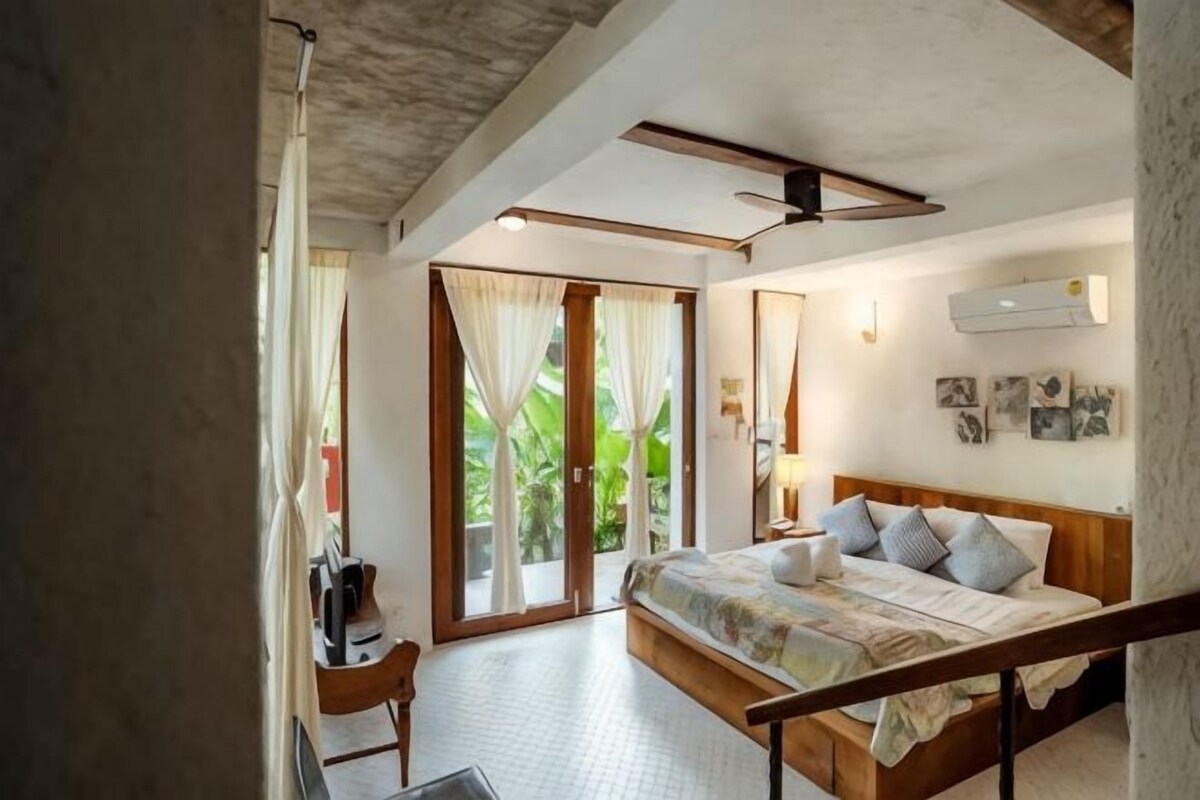 Un dormitorio con cama, escritorio, silla, ventilador de techo y vista a zonas verdes por la puerta.