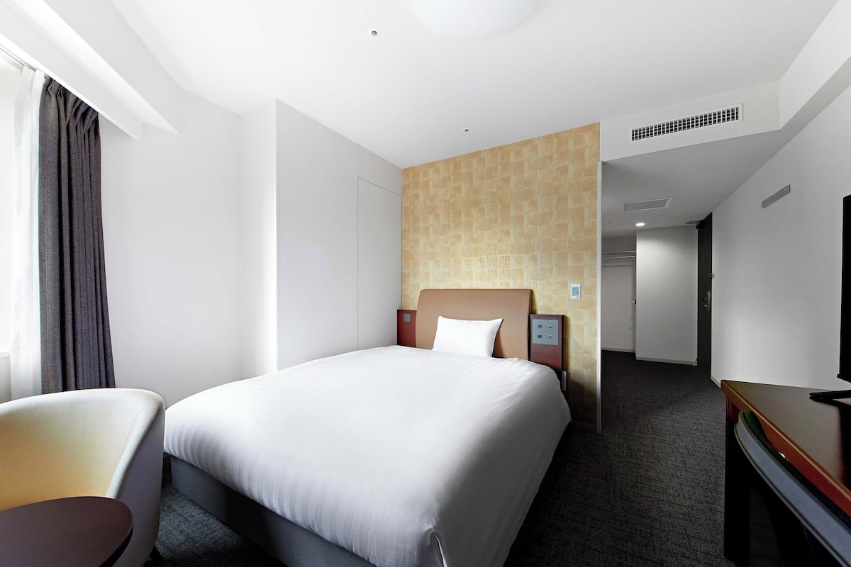 Ein Hotelzimmer mit einem großen Bett, einem Schreibtisch, einem Stuhl und einem Fernseher. Eine Tür führt in einen anderen Raum, und es gibt ein Fenster mit Vorhängen.