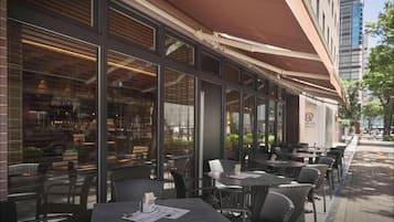 An outdoor seating area with tables and chairs, large windows, and a covered awning.