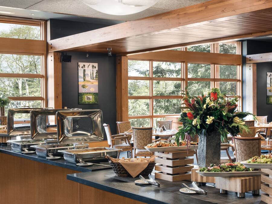 Un buffet avec différents stands de nourriture, un grand vase rempli de fleurs et une vue sur les arbres à travers de grandes fenêtres.
