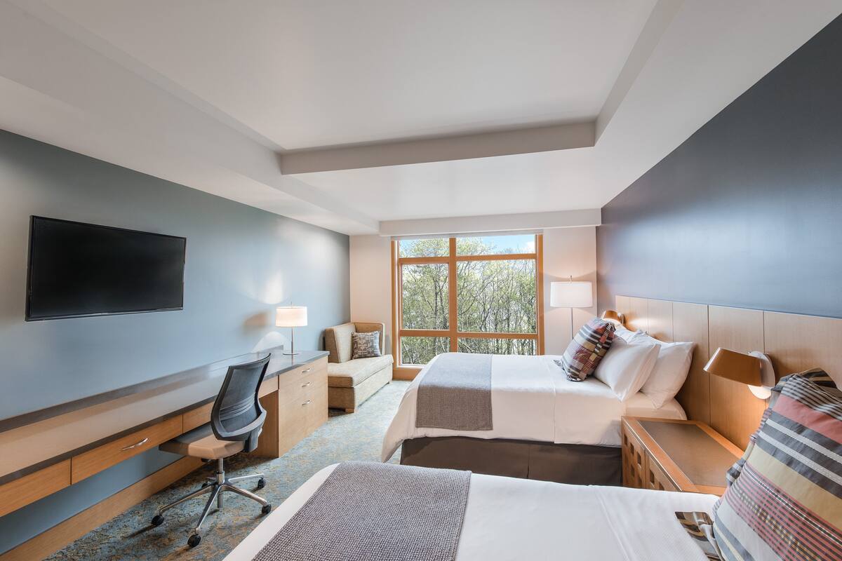 A modern hotel room with a large window, a flat-screen TV, a desk with a chair, and two beds with pillows.