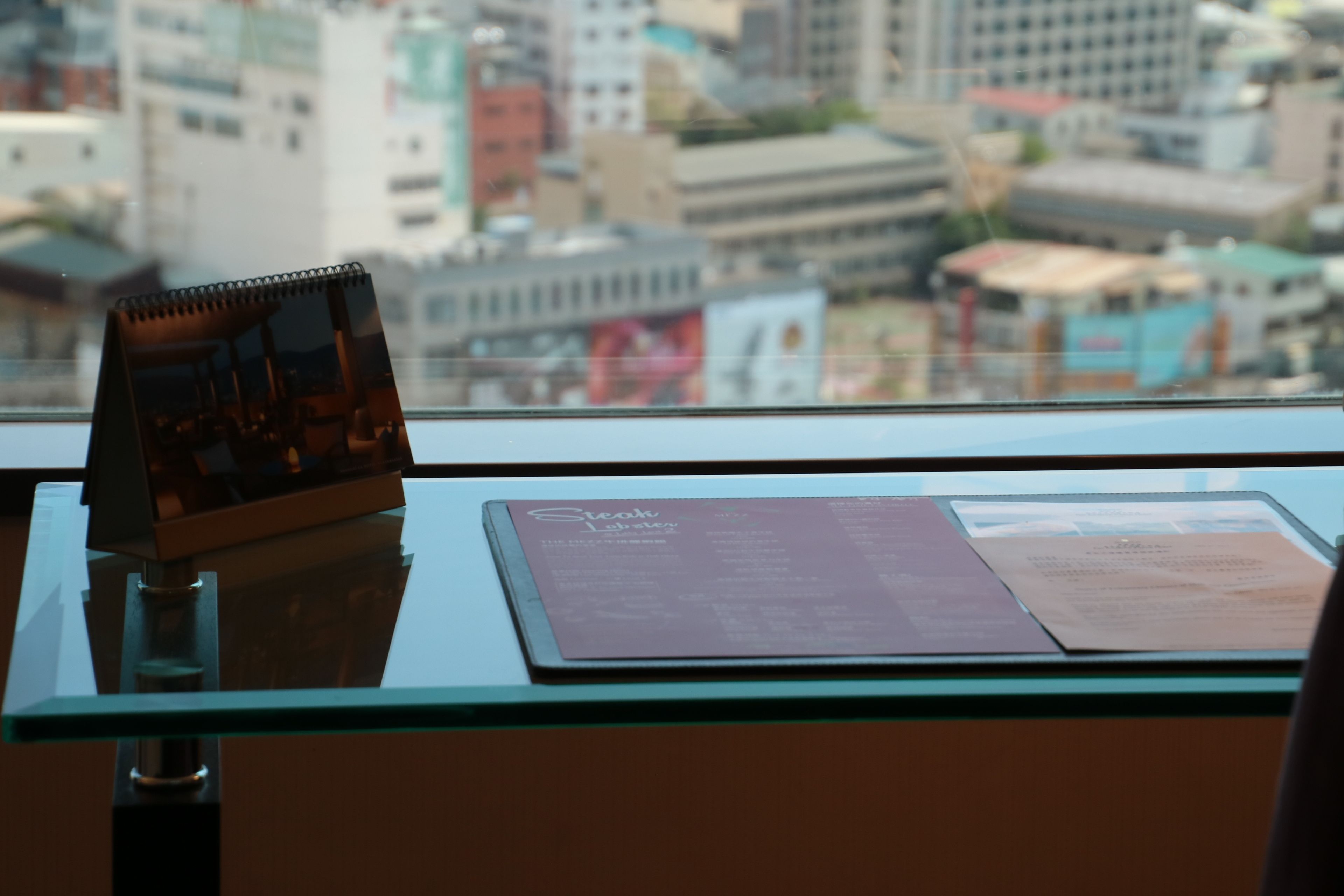 A view from a high-rise window overlooking a cityscape, featuring a glass table with a menu and a notepad.