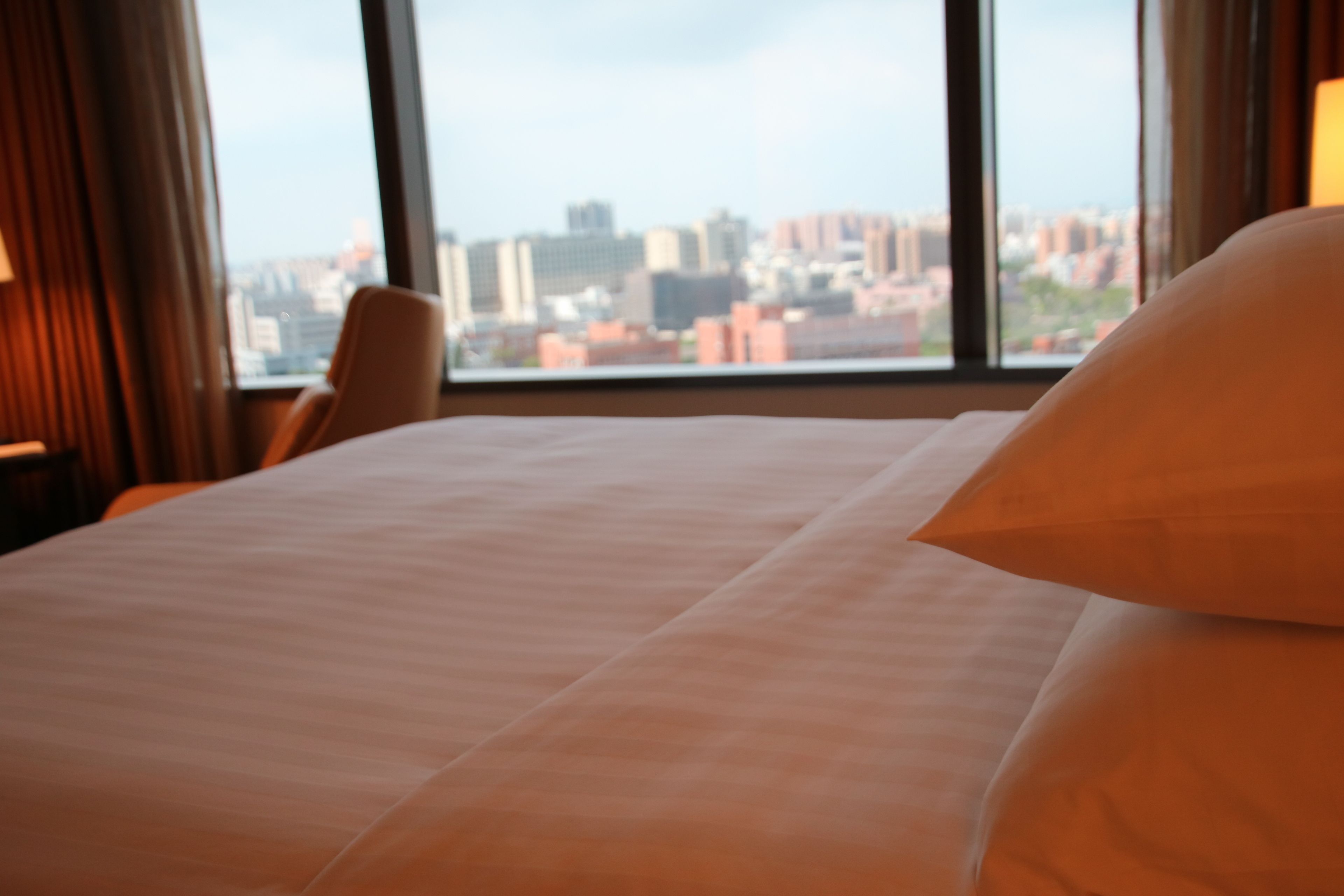 A hotel room with a large bed, a chair, and a view of a cityscape through large windows.
