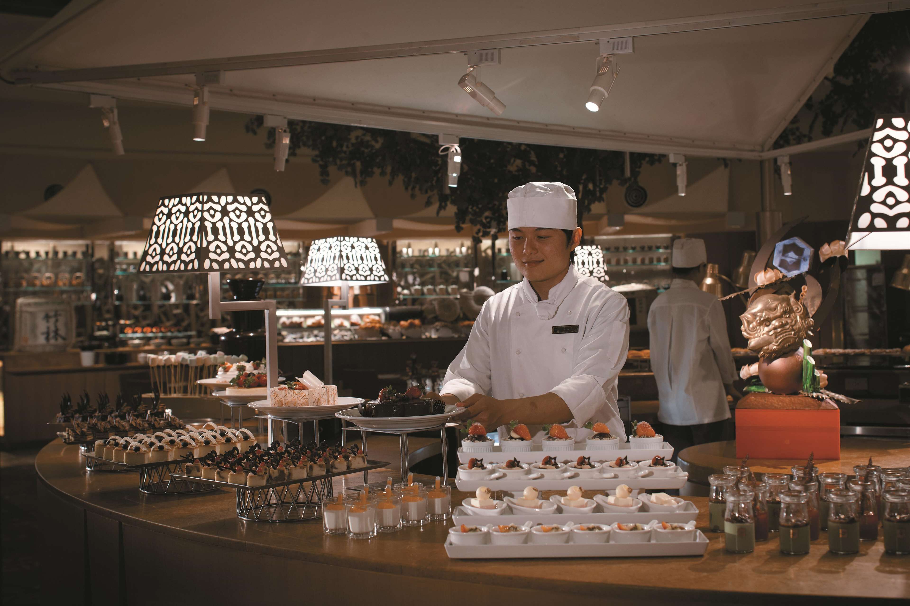A chef in a white uniform preparing food in a well-lit kitchen with various dishes and desserts displayed on the counter.