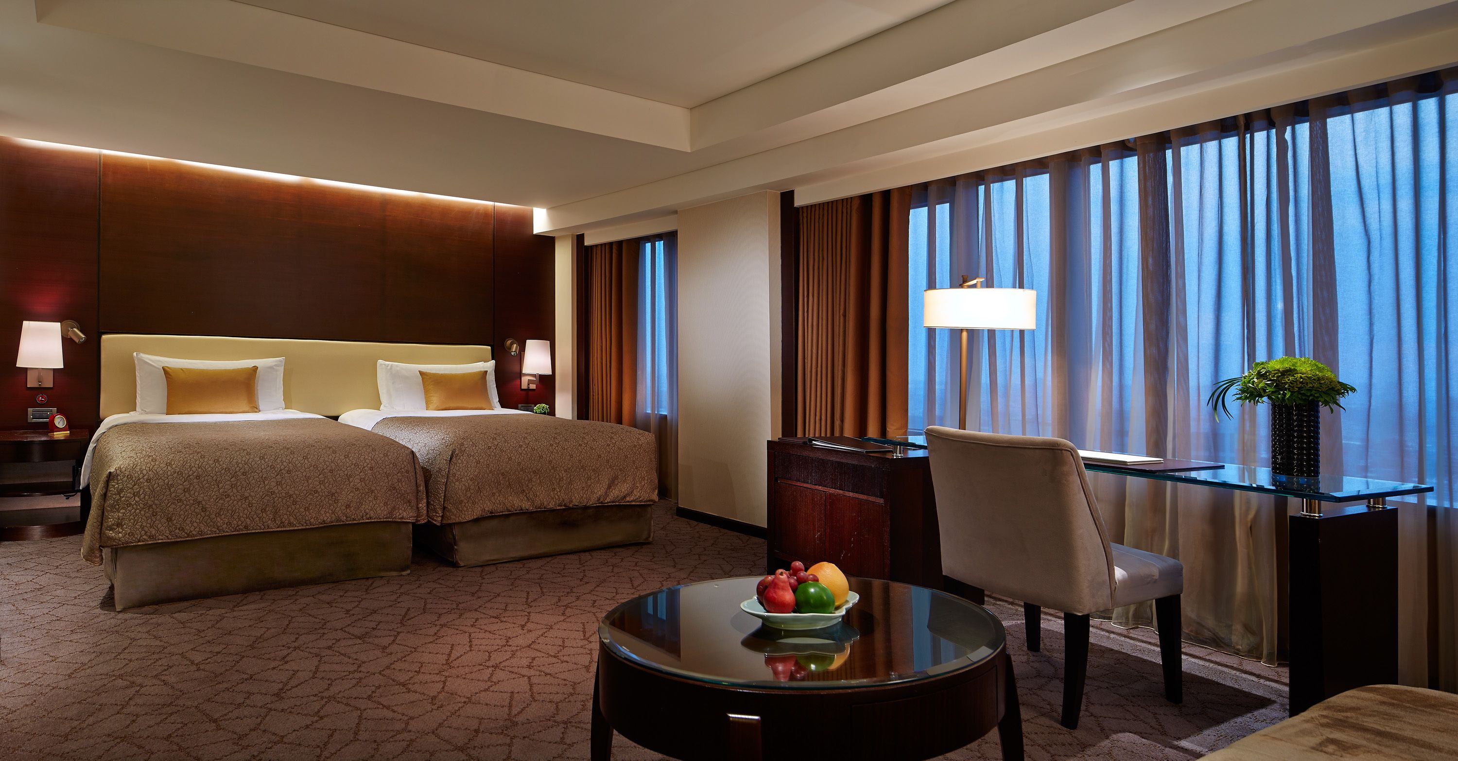 A hotel room with two beds, a desk, a chair, a glass table with fruit, and a large window with curtains.