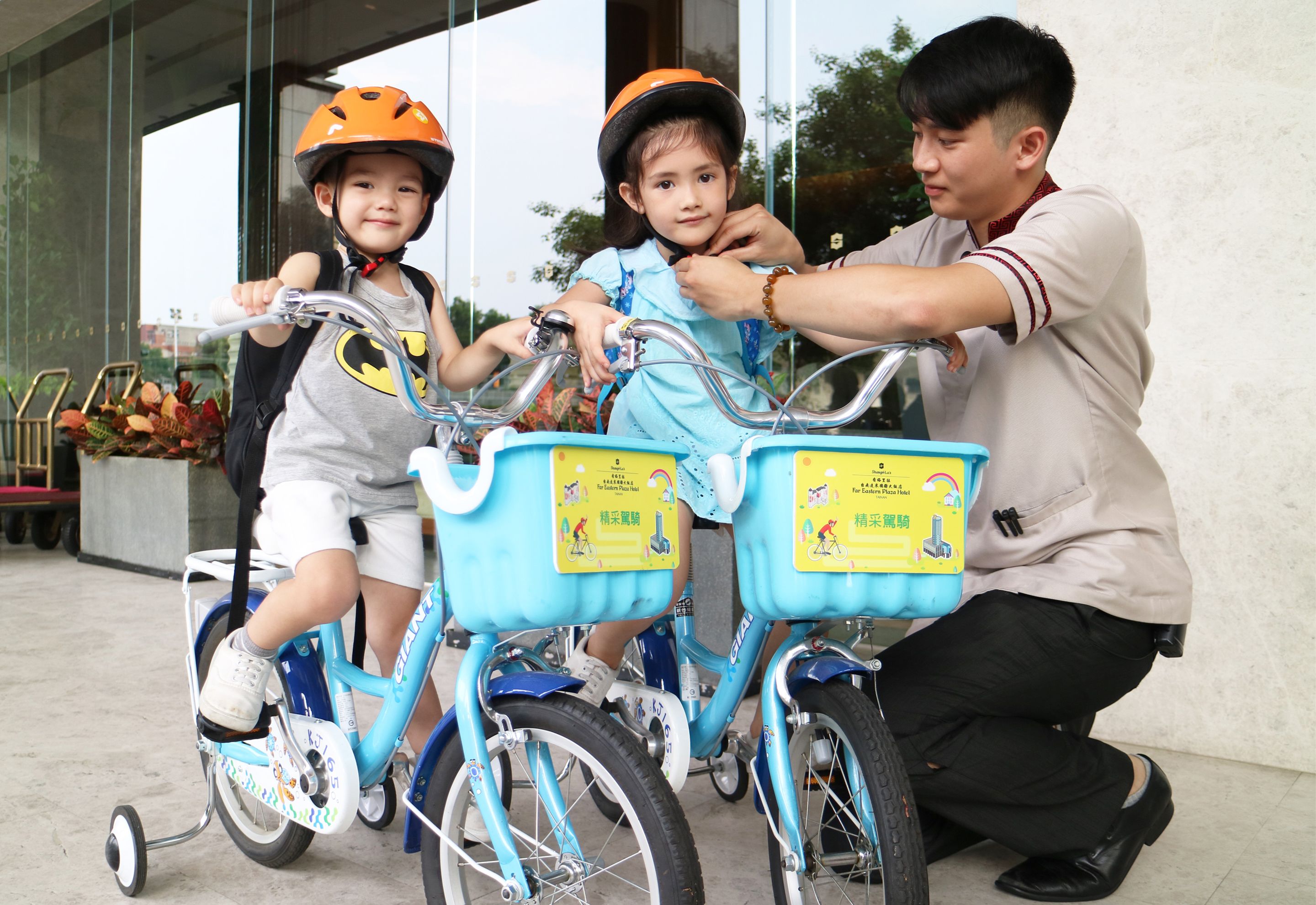 A child on a bicycle with a helmet, an adult assisting, and a building in the background.