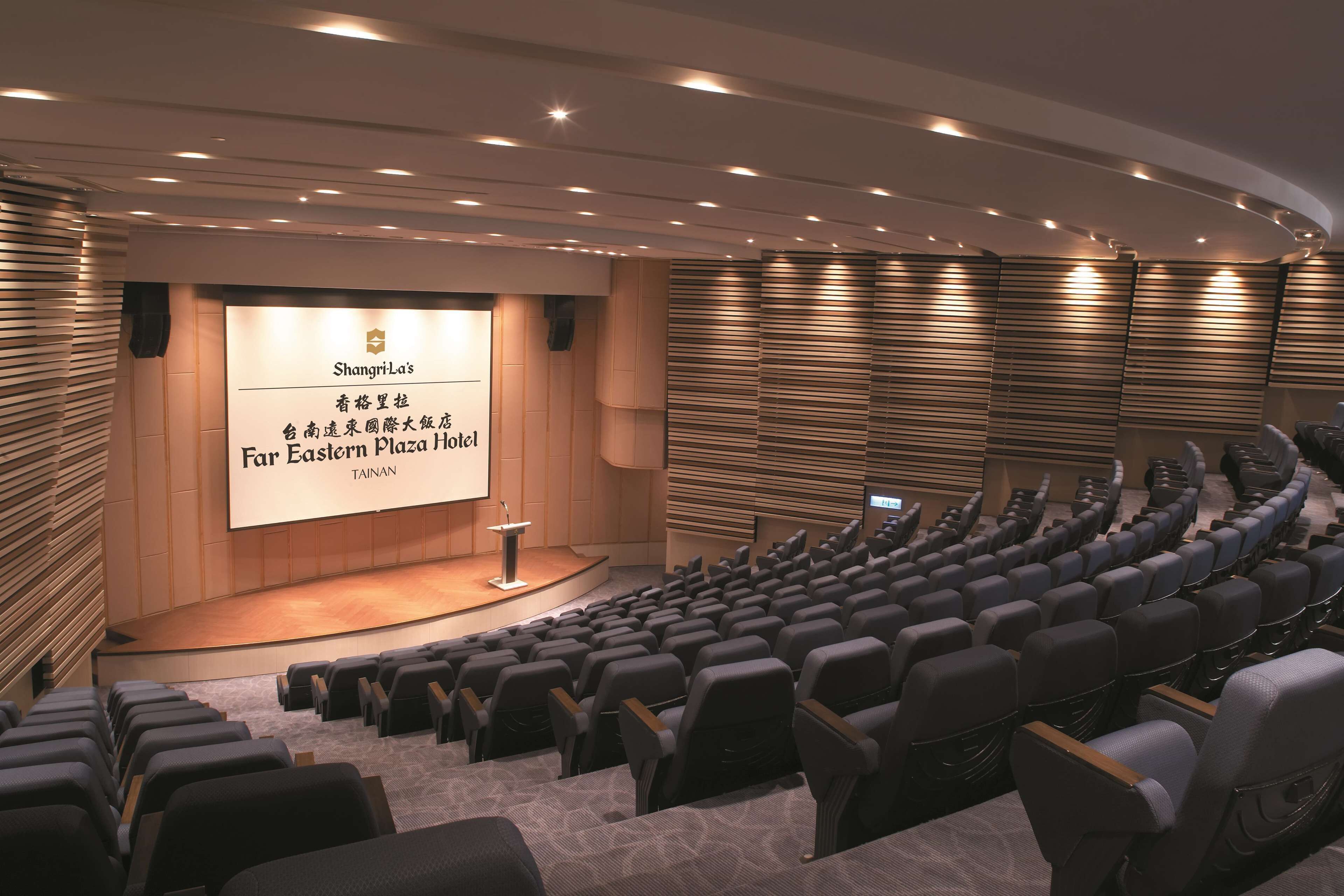 An auditorium with rows of seats, a stage with a podium, and a screen displaying text.