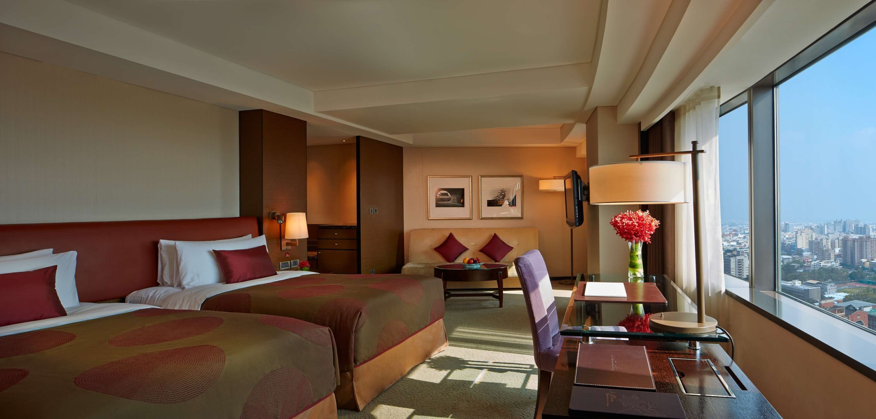 A hotel room with two beds, a desk, a chair, a table, a lamp, a vase with flowers, and a city view through large windows.