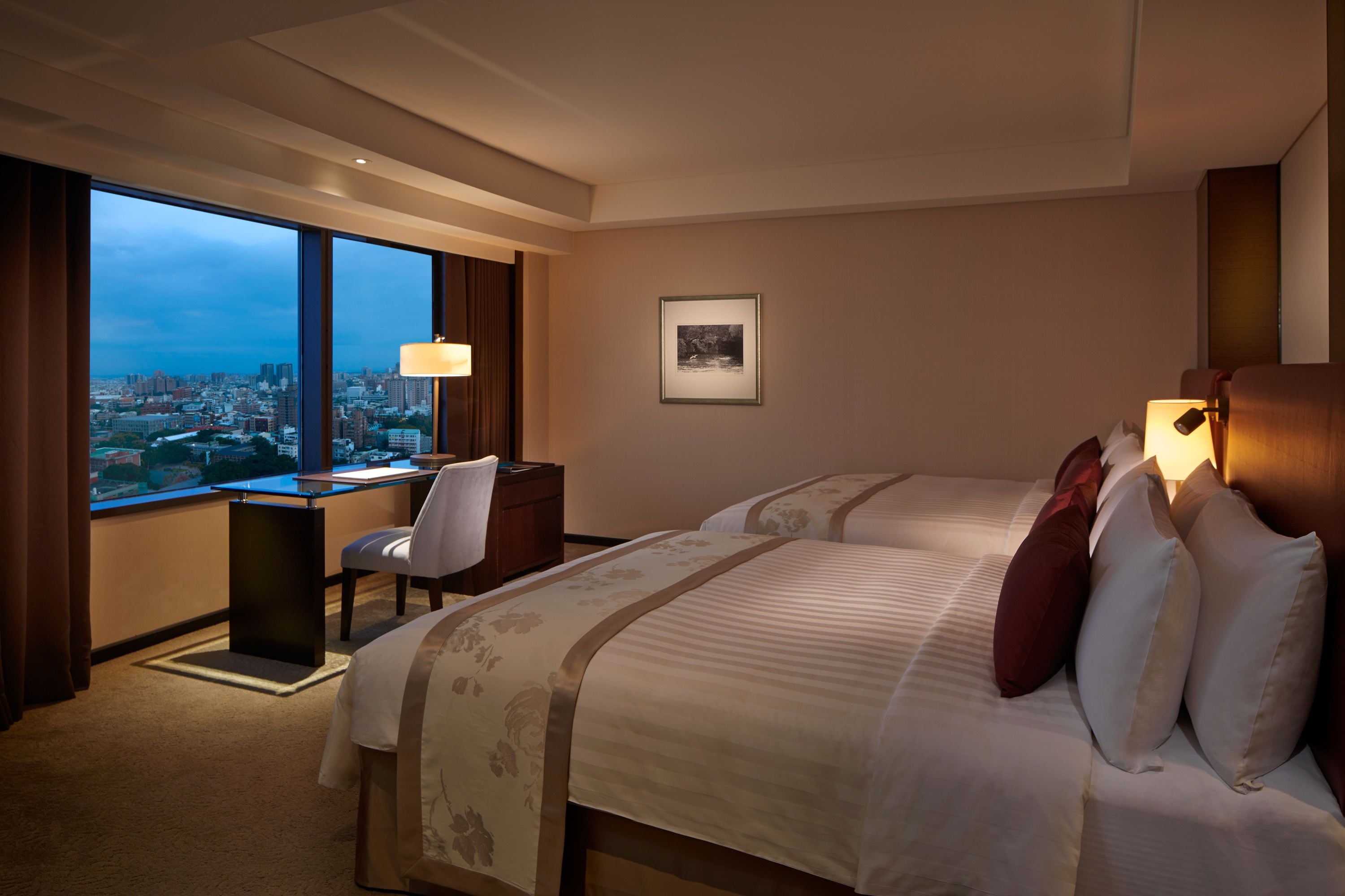 A hotel room with two beds, a desk, and a chair. There is a large window with a city view.