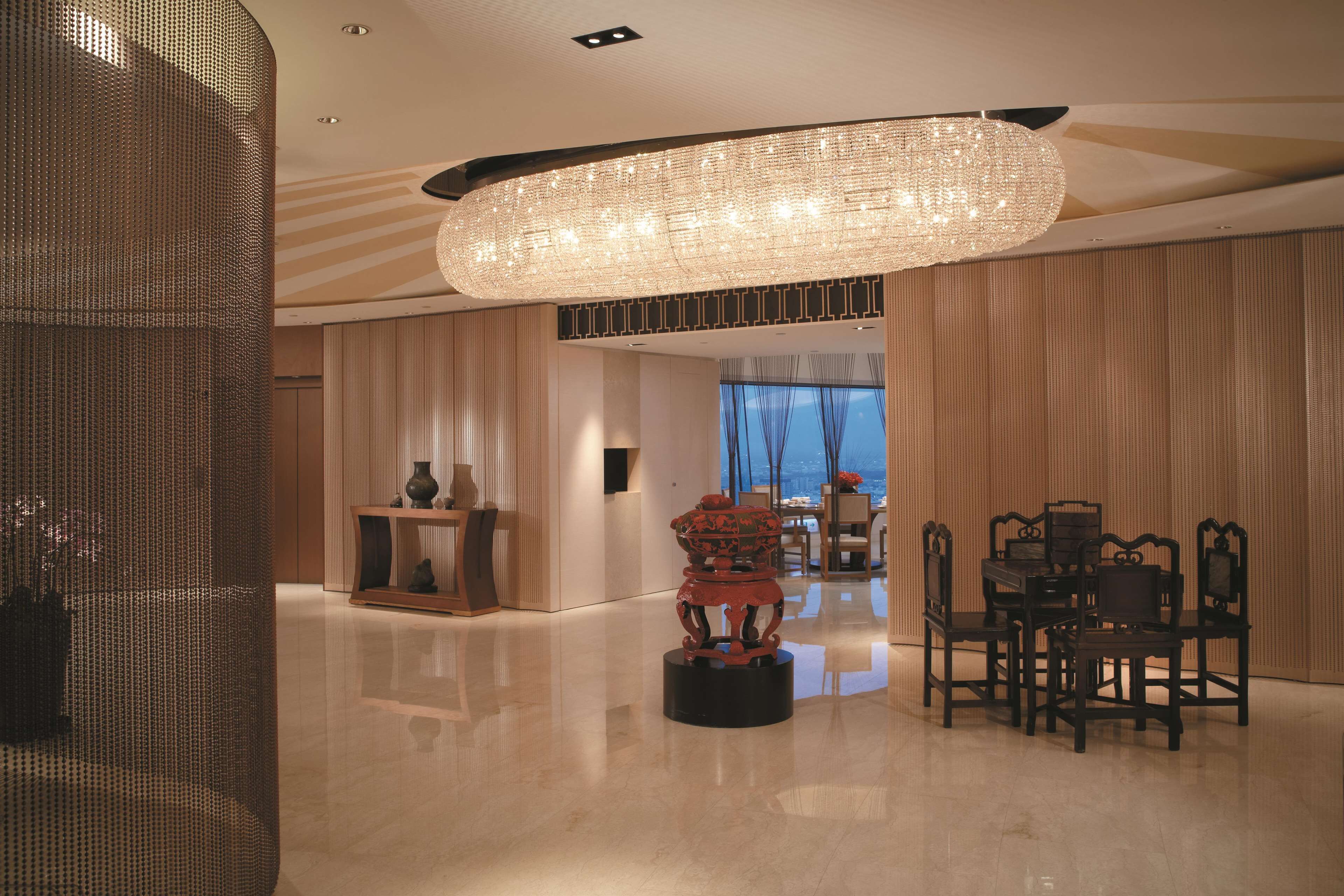 A spacious lobby with a large chandelier, a dining area with wooden furniture, and a view of the cityscape through large windows.