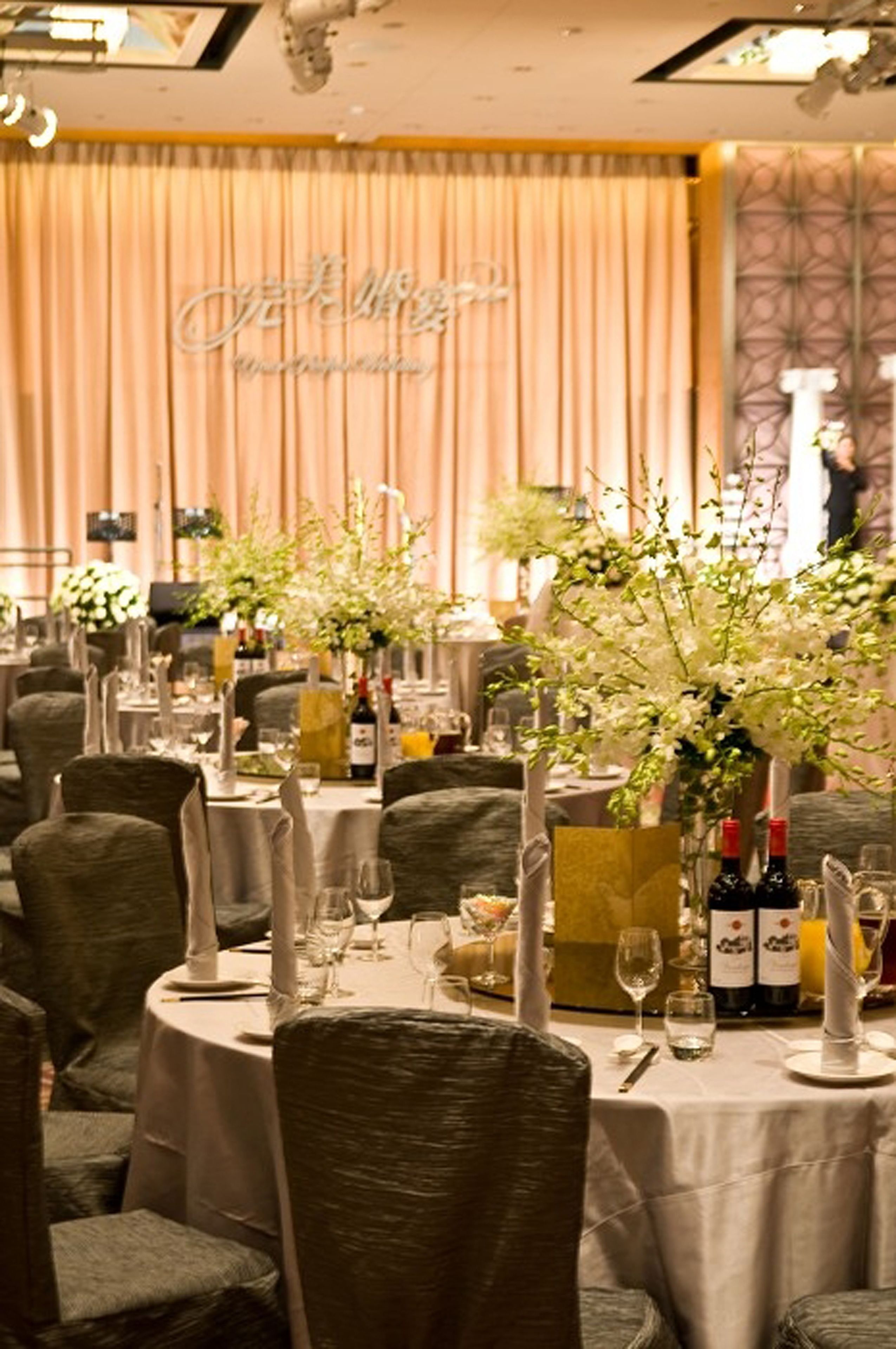 A banquet hall with round tables set for dining, each with a centerpiece of white flowers and candles.