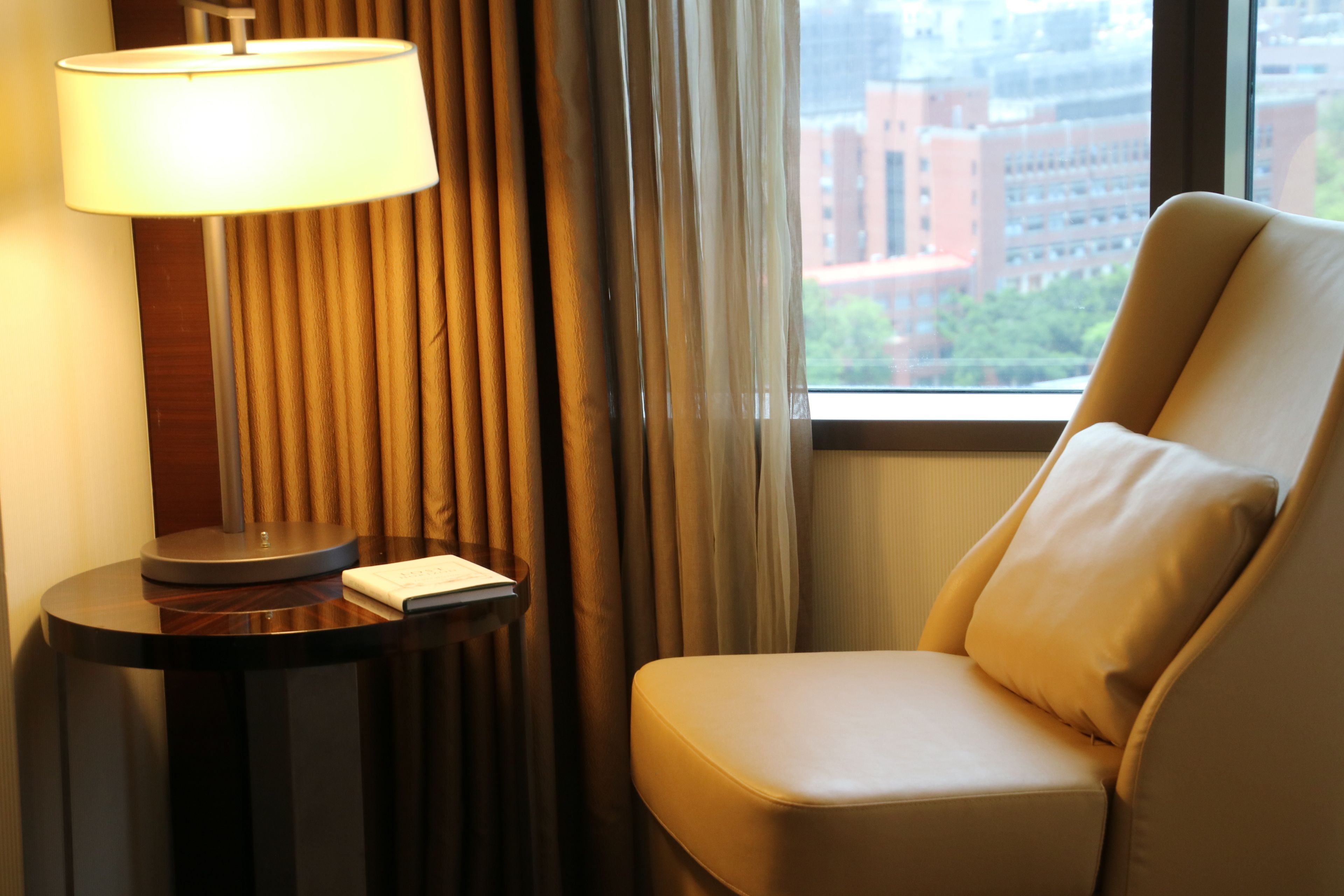A hotel room with a lamp on a side table, a chair, and a view of buildings through the window.
