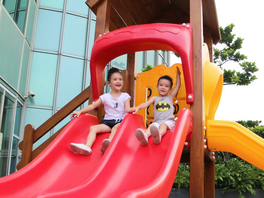 Two children on a red slide at a playground with a wooden structure and a yellow slide.