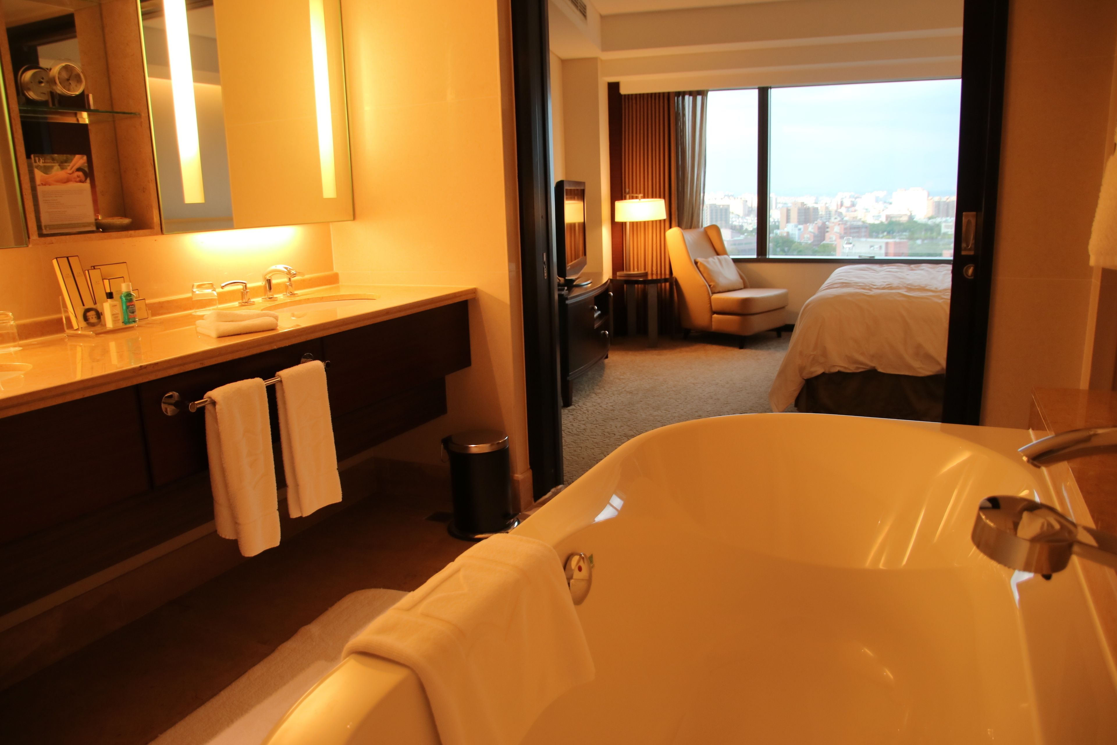 A hotel room with a large bathtub, a vanity with a mirror, a bed, and a view of the city through the window.