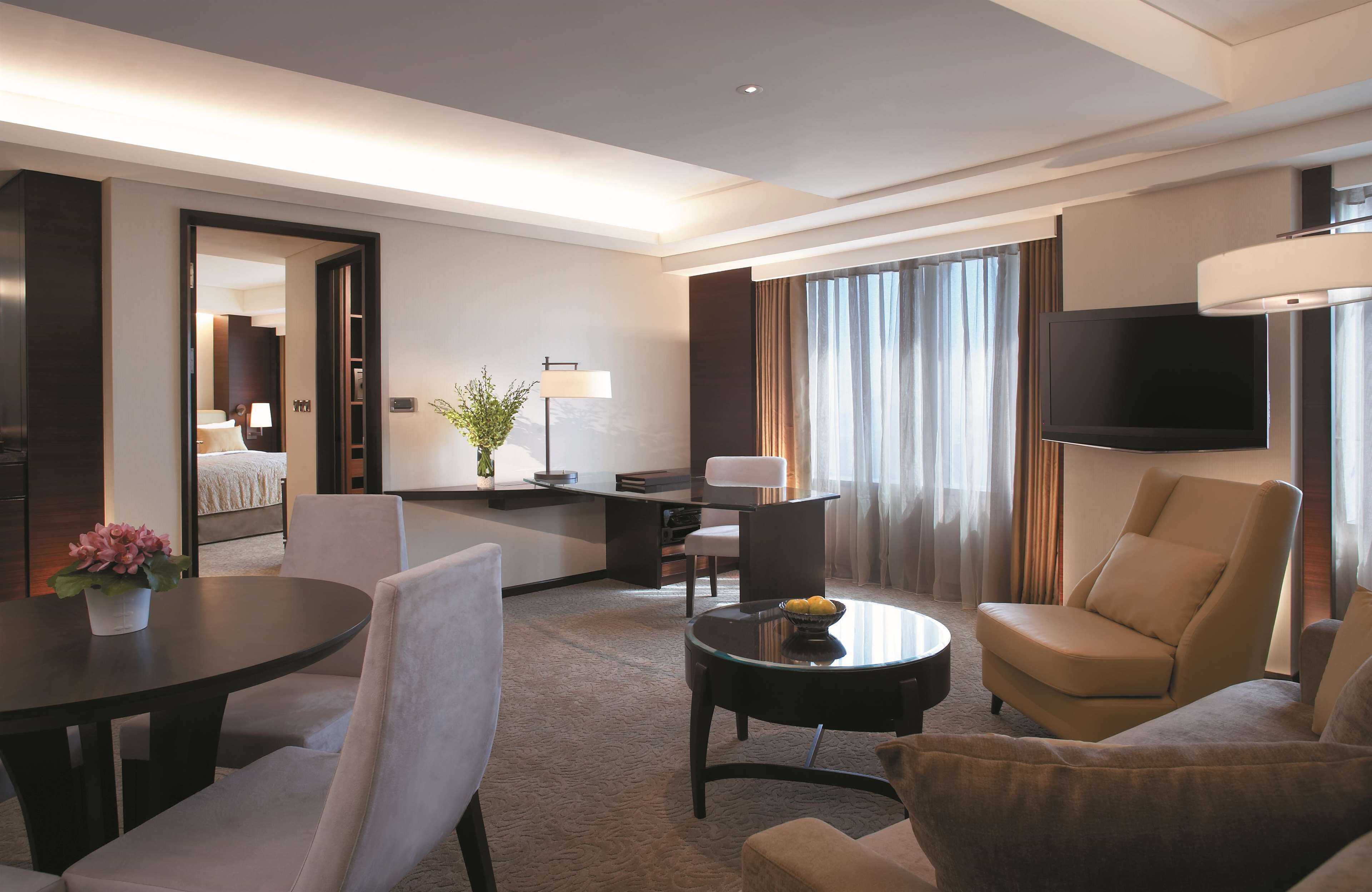 A modern hotel room with a living area featuring a sofa, armchairs, a coffee table, and a TV mounted on the wall.