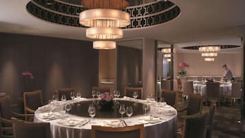 A dining area with round tables set for a meal, a central chandelier, and a person in the background.