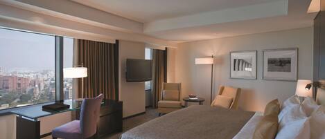 A hotel room with a large bed, a desk, a chair, a TV, and a view of the city.