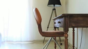 Een stoel met een hoge rugleuning en metalen poten, een houten tafel met lades, en een vloerlamp in een kamer met een lichtgekleurde vloer en wanden.