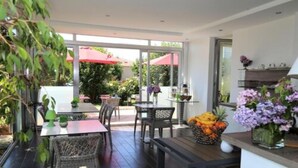 An indoor patio area with tables and chairs, a small kitchen area with a fruit bowl, and a view of the outside garden.