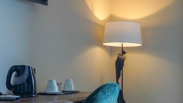 A hotel room with a desk, a teal chair, a television, a coffee maker, a lamp, and an air conditioner.