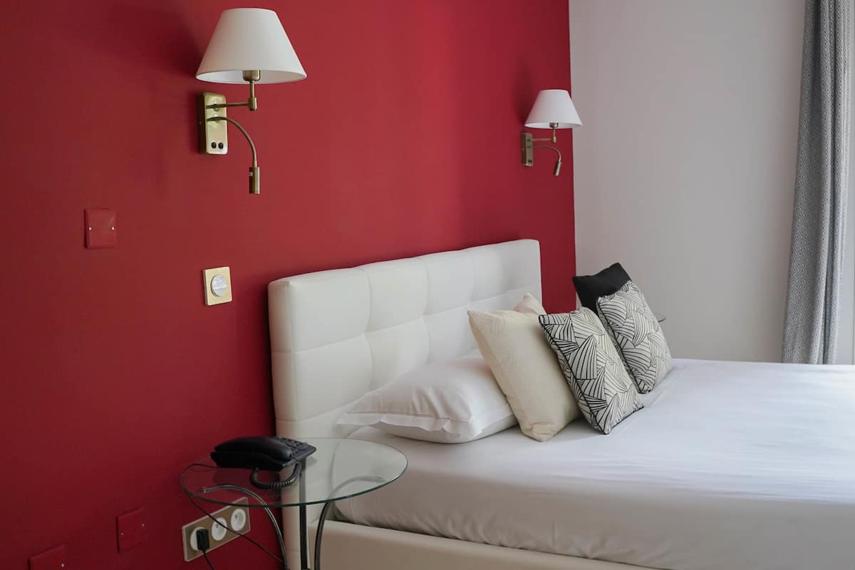 Ein Hotelzimmer mit roter Wand, einem weißen Bett, zwei Wandlampen, einem Nachttisch mit Telefon und einem Fenster mit Blick auf Grünflächen.