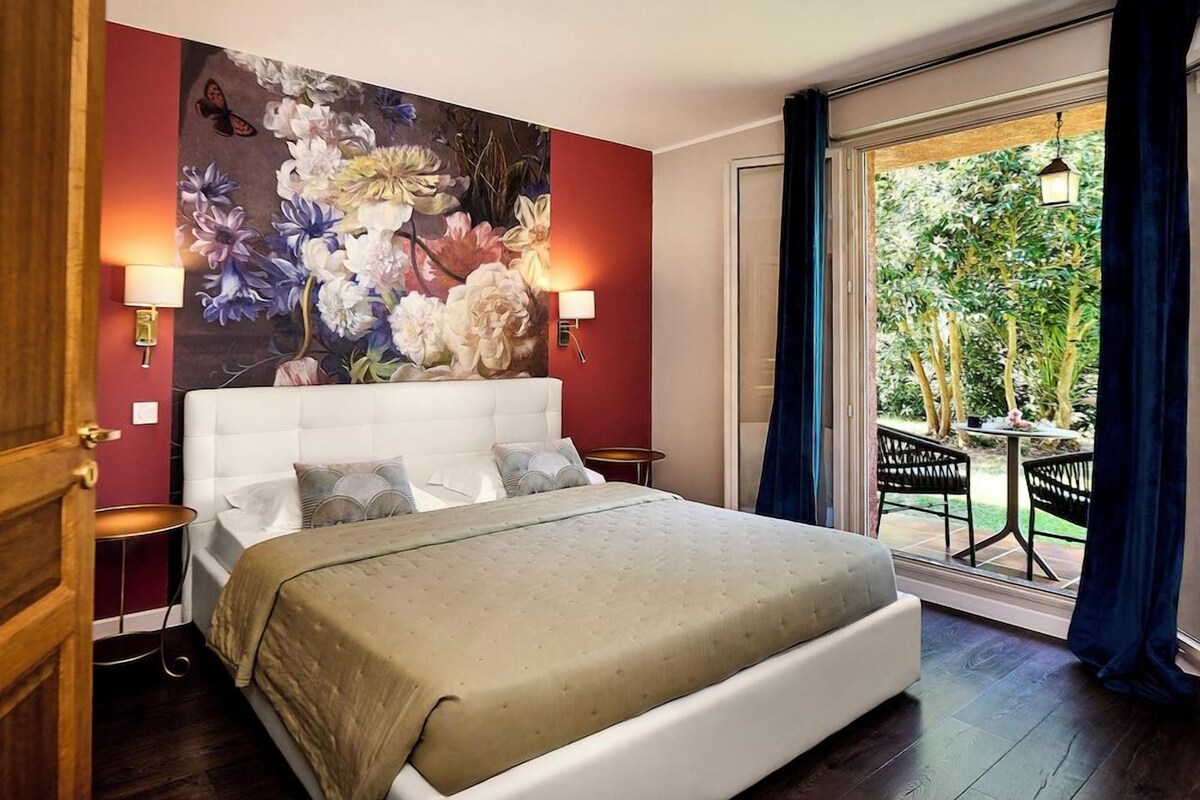 A bedroom with a large bed, a floral wall mural, two bedside lamps, and a view of an outdoor garden through a sliding door.