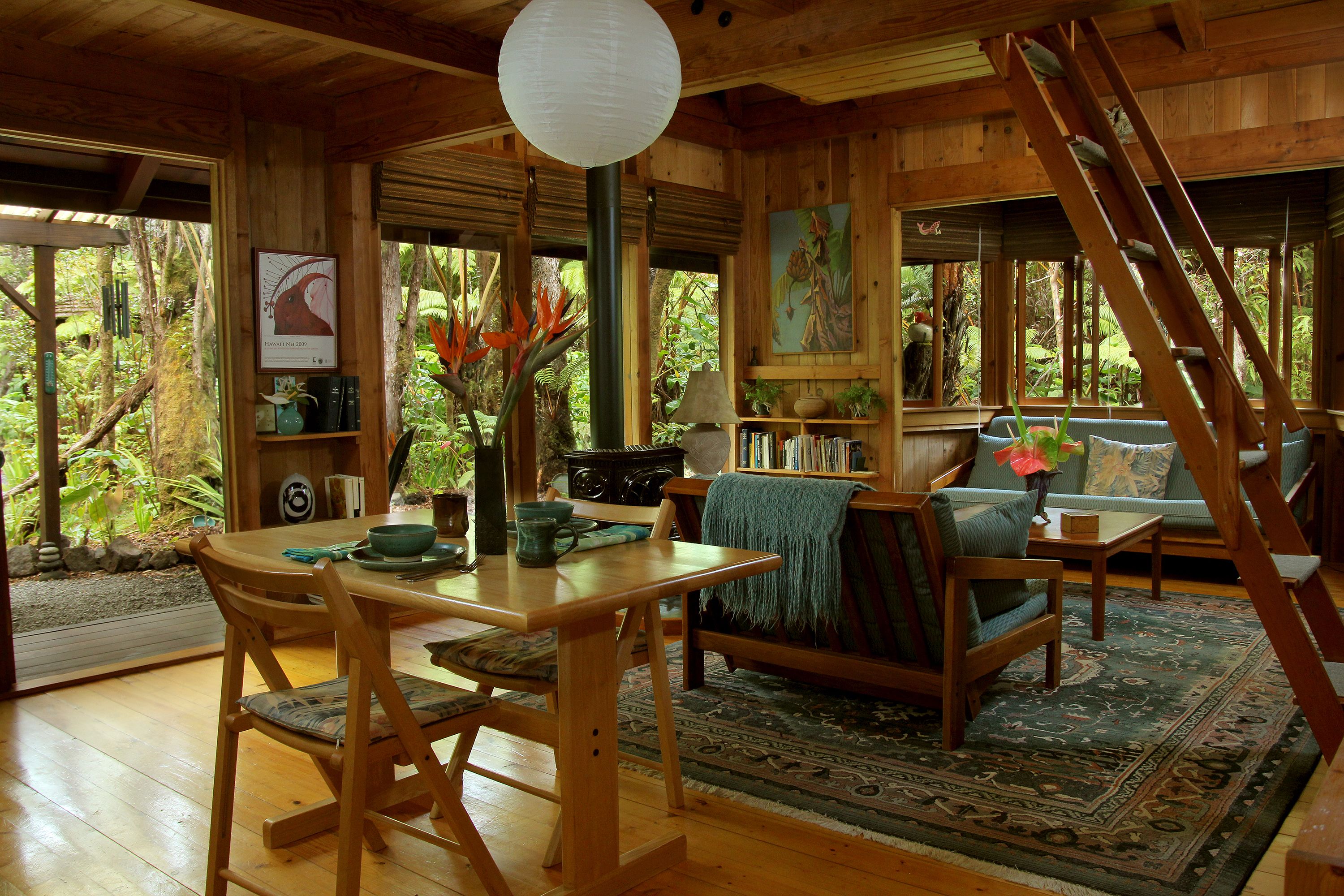 A wooden interior with a dining table, chairs, a sofa, a ladder, and a view of the surrounding forest.