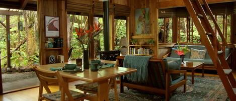 A wooden interior with a dining table, chairs, a sofa, a ladder, and a view of the surrounding forest.