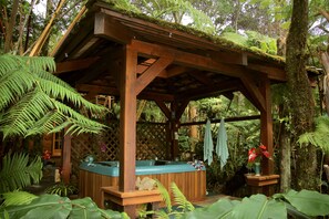 A wooden gazebo with a hot tub, surrounded by lush greenery and ferns.