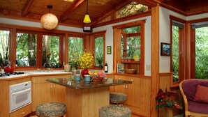 A kitchen with wooden cabinets, a central island with stools, and large windows with tropical views. - Volcano Rainforest Retreat (Volcano)