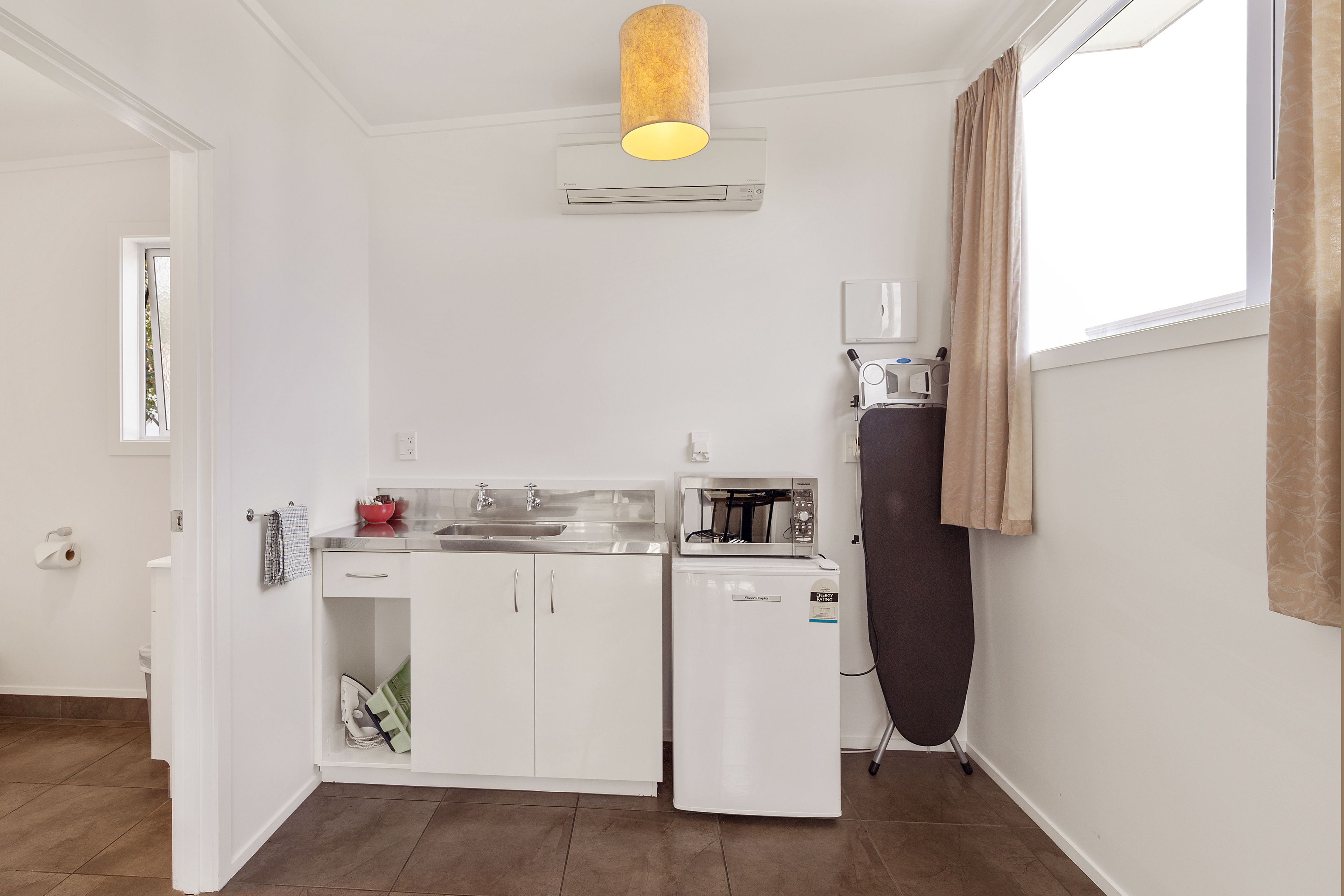 Cocina compacta con armarios blancos, microondas integrado, un refrigerador pequeño y una tetera.