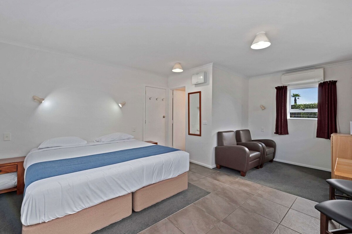 Une chambre d’hôtel avec un lit, deux chaises, un bureau et une fenêtre avec des rideaux.