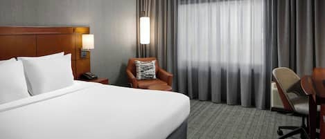A hotel room with a large bed, a nightstand with a lamp, a chair, and a desk.