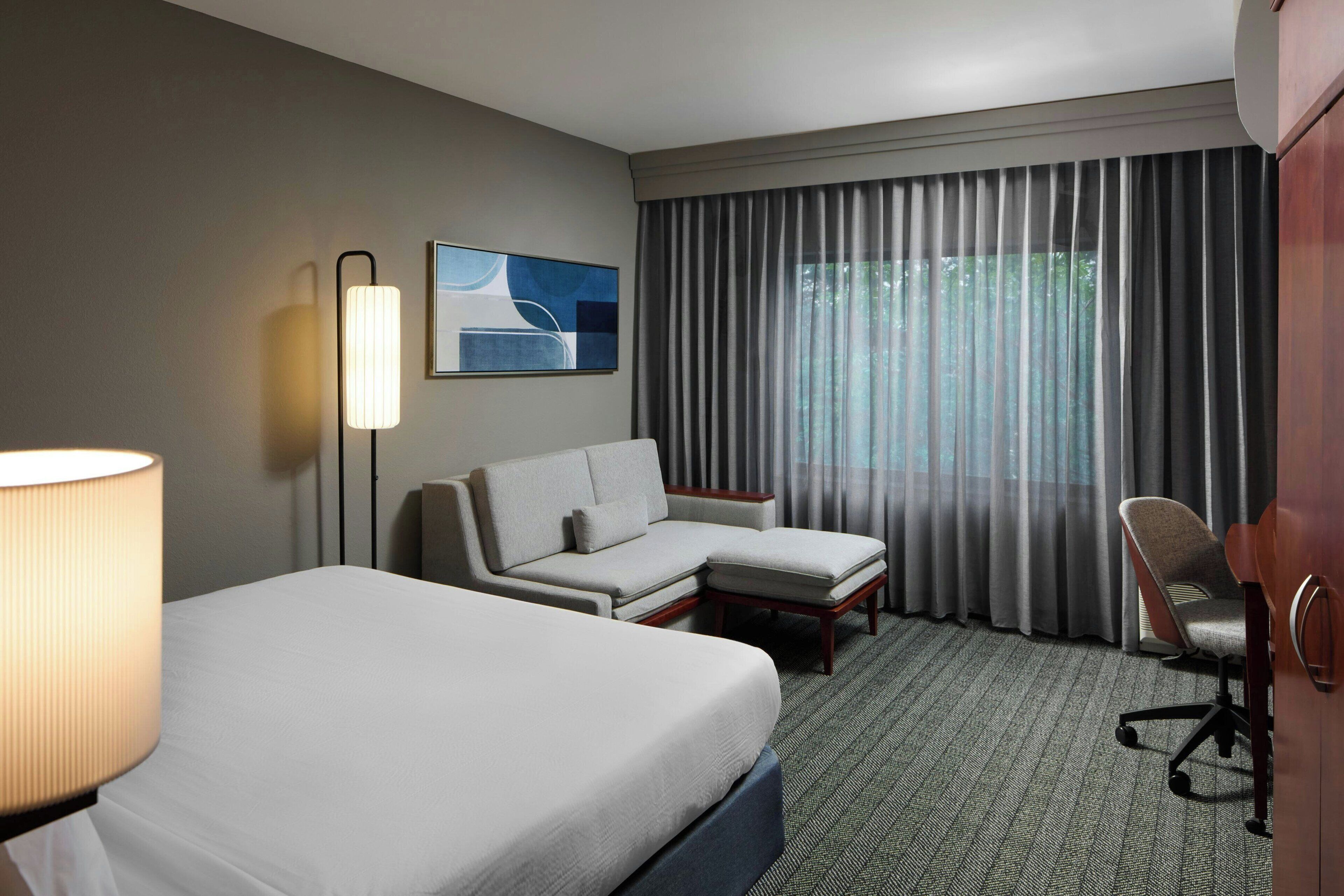 Una habitación de hotel con una cama grande, un sofá, una silla, un escritorio, una lámpara, un cuadro y una ventana con cortinas.