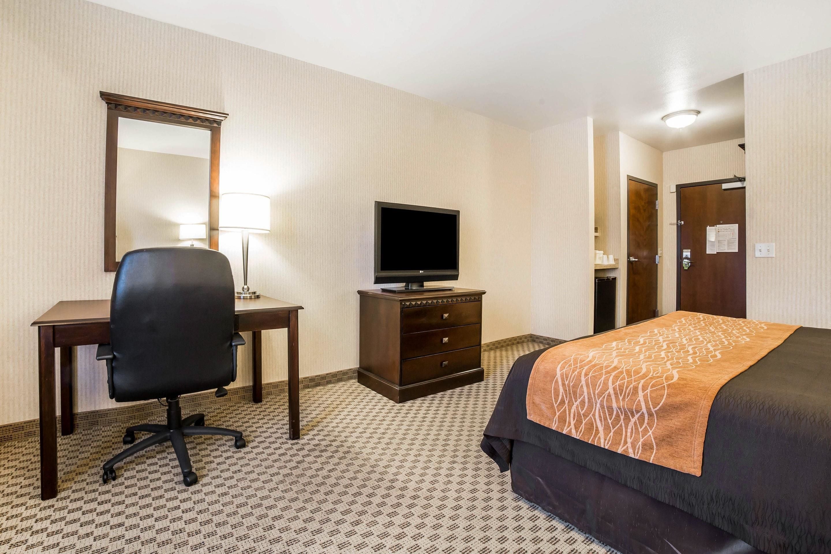 A hotel room with a bed, a desk with a chair, a television, a dresser, and a mirror.