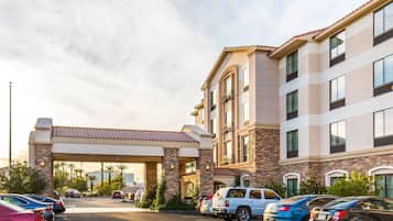 Un hotel con entrada cubierta, varios espacios de estacionamiento y un edificio de fachada de piedra y numerosas ventanas.